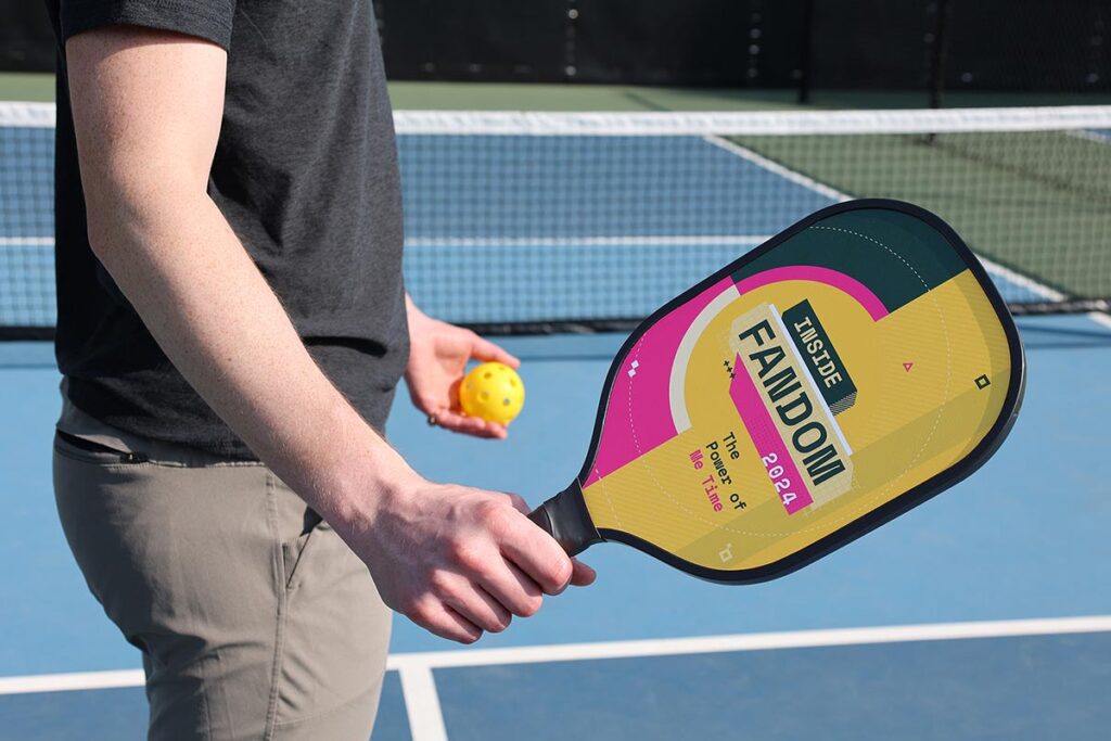 A person holds a pickleball paddle with a colorful design featuring the words "TABE FANDOM 2024" and "There is Joy in the Journey." They also hold a yellow pickleball while standing on a court.