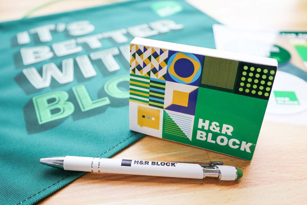 A green and white pen and a colorful card with geometric patterns are placed on a table. Both items display the H&R Block logo. In the background, there's a green cloth with the text "It's Better With Block.