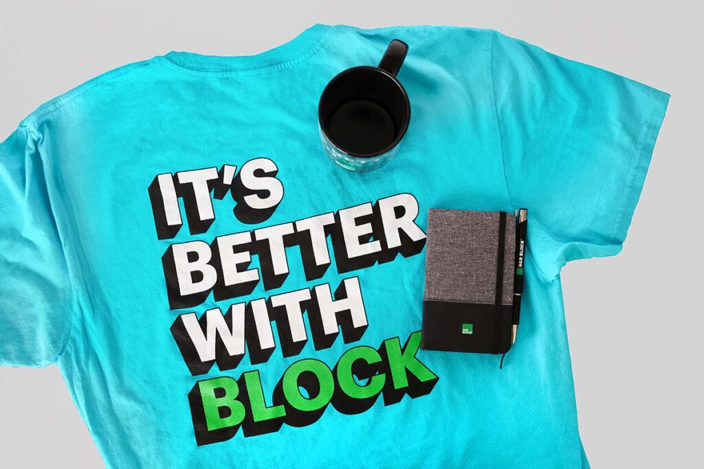 A turquoise T-shirt with the text "IT'S BETTER WITH BLOCK" in bold white and green letters. Next to the shirt, there is a black mug containing coffee and a small notebook with a pen on top.