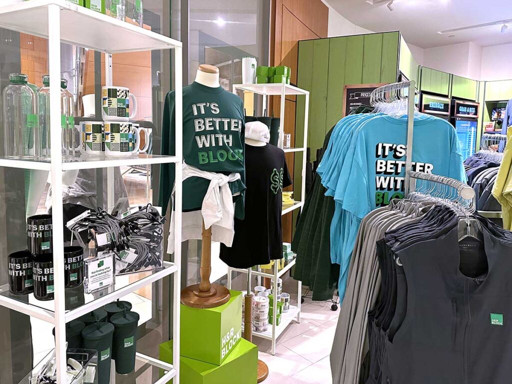 A clothing store display featuring mannequins wearing green and blue shirts that read "It's Better with Block." Shelves hold water bottles, mugs, and various accessories. Racks of similar shirts and apparel surround the area.