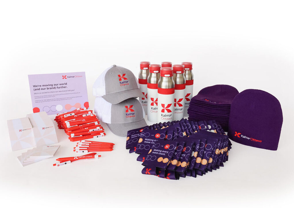 A collection of branded merchandise including water bottles, caps, beanies, lanyards, pens, and pamphlets. Items are organized on a plain background, prominently featuring the Kantar brand logo in red and purple.