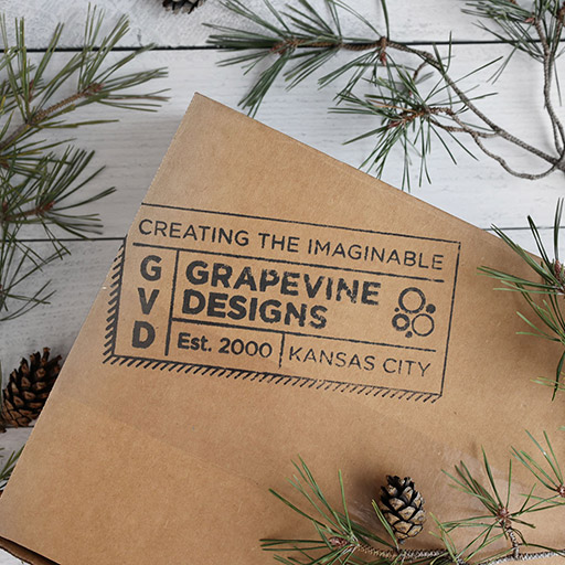 A cardboard package from Grapevine Designs labeled "Creating the Imaginable" and "Est. 2000, Kansas City" is surrounded by pine branches and pinecones on a white wooden surface.