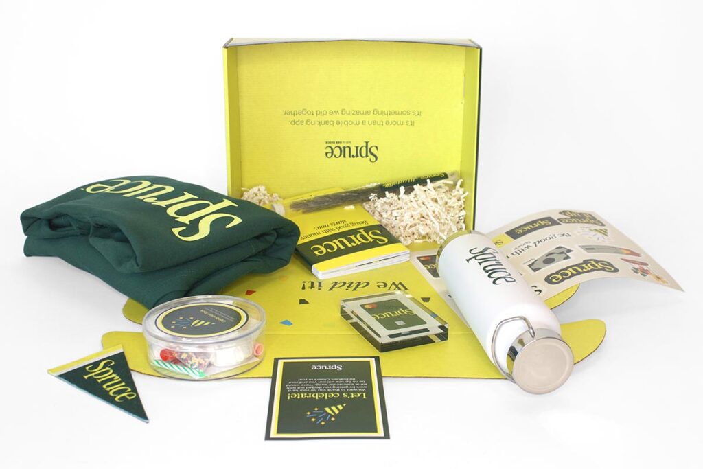 A yellow box labeled "Spruce" containing a green shirt, a white bottle, a packed tin, brochures, and small promotional materials is displayed. The box is partially filled with shredded paper for packing.