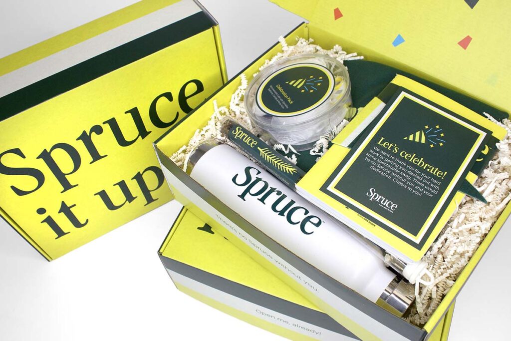 A bright, festive gift box with "Spruce" on a water bottle, a pen, a candle, and a card that says "Let's Celebrate!" The box is filled with crinkled paper for decoration. The lid reads "Spruce it up" in bold letters.