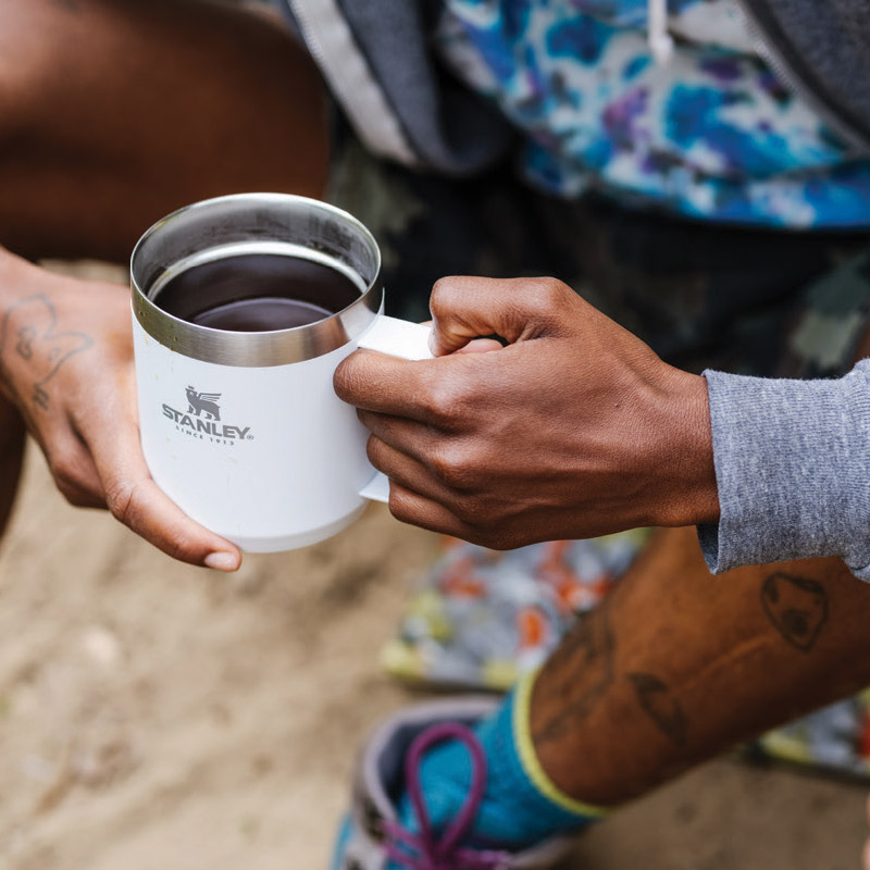 A person holding a white Stanley mug filled with a dark beverage. They are wearing colorful shorts and patterned socks, and sitting outdoors on a sandy surface. Tattoos are visible on the person's arms and legs.