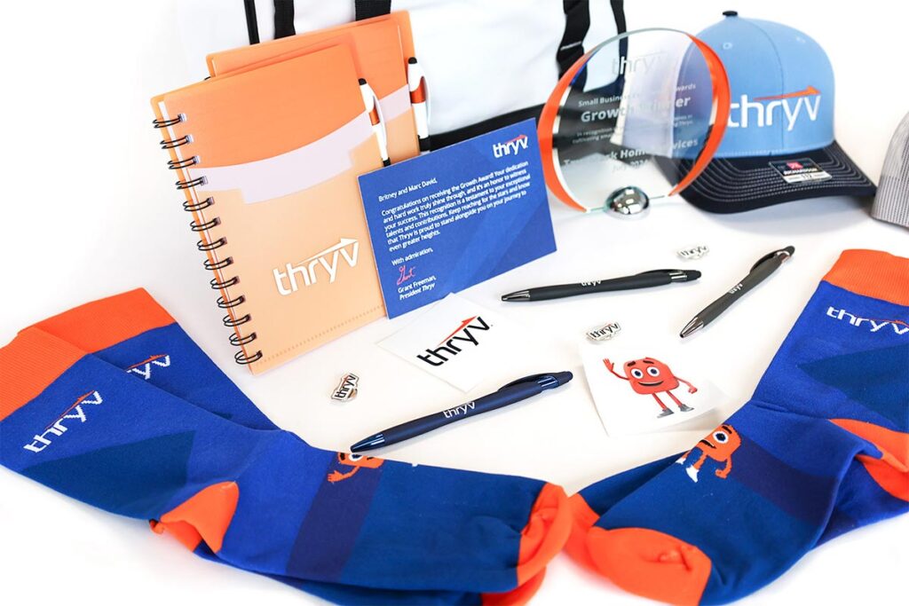 A collection of promotional items including a spiral notebook, pens, a cap, socks, stickers, and a card all featuring the Thryv logo. An award plaque is also displayed amidst the items. The color scheme includes blue, orange, and black.