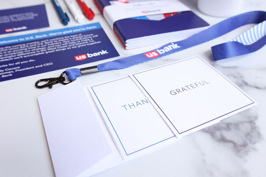 A close-up image featuring a lanyard with a badge holder, a note with "Thankful" and "Grateful," and US Bank branded materials, including pamphlets and a pen, on a marble surface.