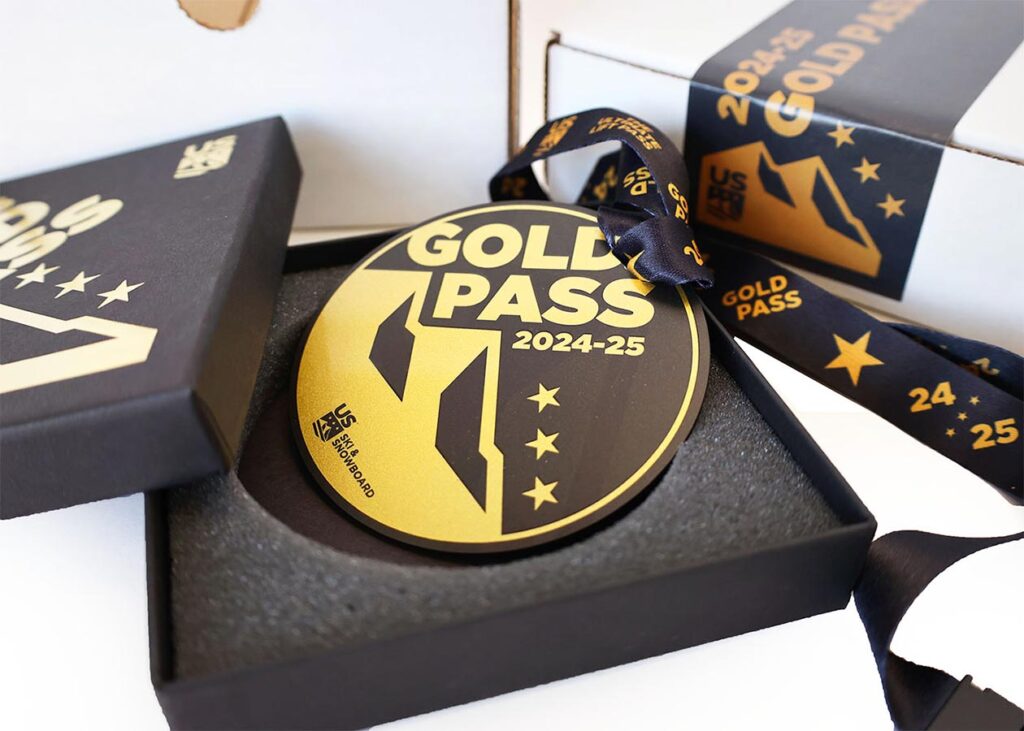 A gold pass for 2024-25 sits in a black box with a gray interior. The pass features a mountain logo, stars, and text "US Ski and Snowboard." A matching lanyard is attached.