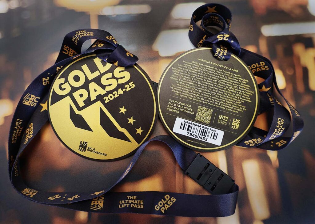 Two round gold ski passes with "GOLD PASS 2024-25" written on them, attached to dark lanyards with text. The back of one pass shows usage instructions, a QR code, and a barcode. The lanyards read "THE ULTIMATE LIFT PASS.