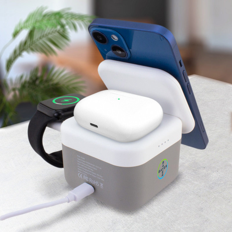 A multi-device charging station on a table with a smartphone, smartwatch, and wireless earbuds charging simultaneously. The setup is against a blurred background with some green plant leaves.