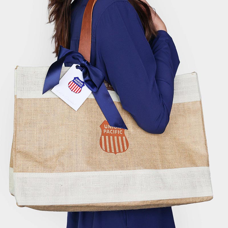 A person in a blue outfit carrying a beige tote bag with a brown Union Pacific logo. The bag has white accents and a dark blue ribbon attached to the handle with a Union Pacific tag.