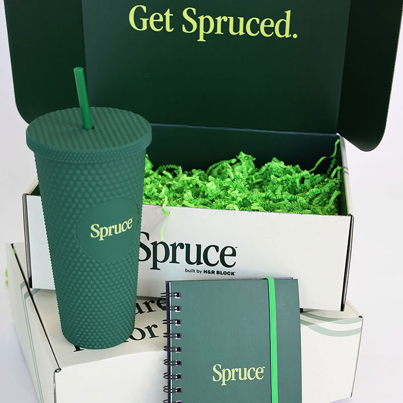 A green cup with a textured surface and a straw, a notebook, and a box filled with shredded paper, all featuring the logo "Spruce". The box is partially closed and displays the text "Get Spruced.