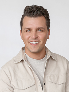 A smiling man with short dark hair is wearing a beige shirt over a light gray t-shirt. The background is plain white.