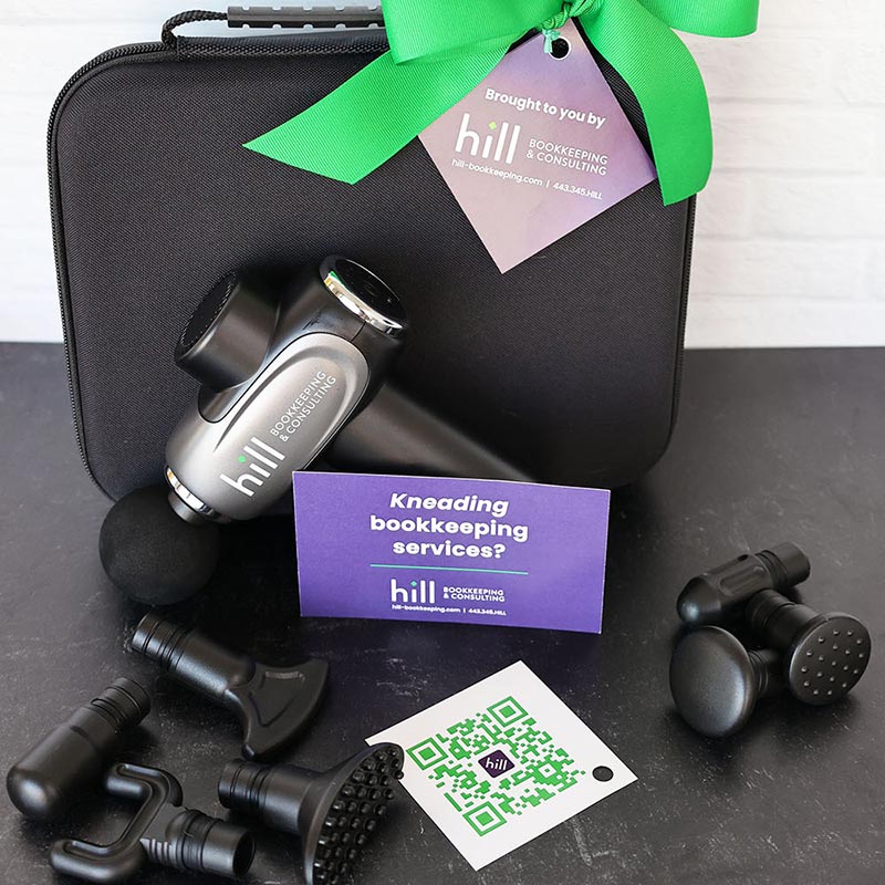 A massage gun set with multiple attachments placed on a black case. A card reads "Kneading bookkeeping services?" A green ribbon with a tag shows "Hill Bookkeeping & Consulting" alongside QR code and contact details.