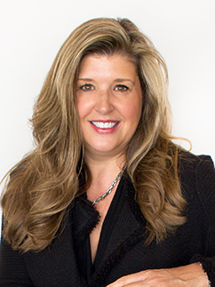 A woman with long, wavy blonde hair smiles at the camera. She is wearing a black jacket and a silver necklace, standing against a plain white background.