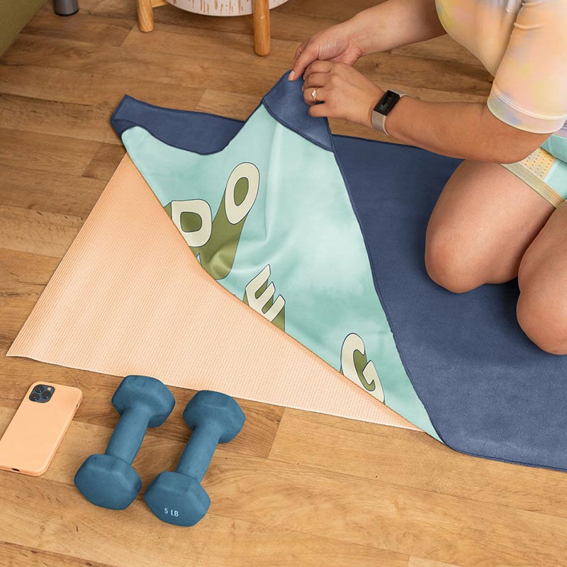 A person kneels on a wooden floor, folding a yoga mat with a colorful design. Two teal dumbbells and a smartphone are placed nearby. The individual wears a smartwatch and light-colored clothing.