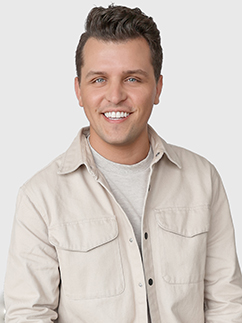 Smiling person with short dark hair wearing a light-colored jacket over a gray shirt, looking at the camera against a plain background.