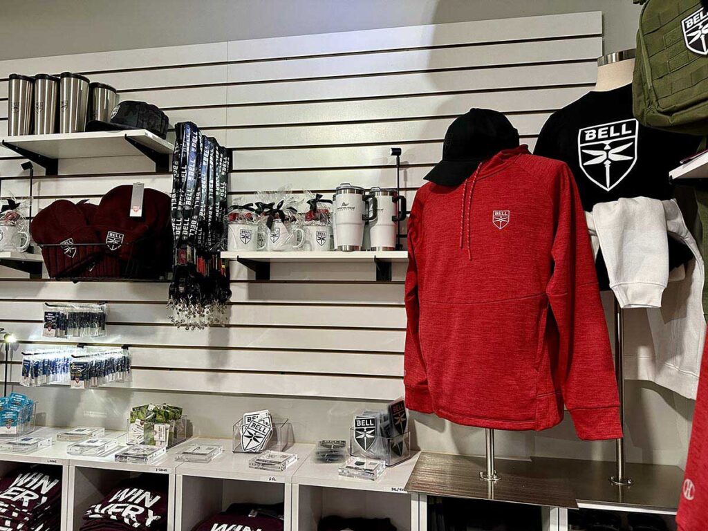 A retail display featuring branded merchandise: red hoodies, black and white shirts, scarves, mugs, and travel tumblers with a logo. Items are neatly arranged on shelves and racks against a white slatted wall.