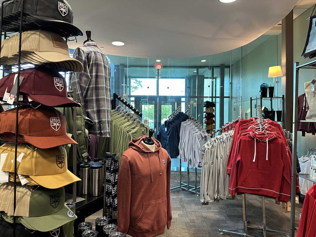 A retail clothing store displays various apparel. Hats and mugs are on the left, while hoodies in orange, blue, and red are on racks. A glass entrance with trees visible outside is in the background.