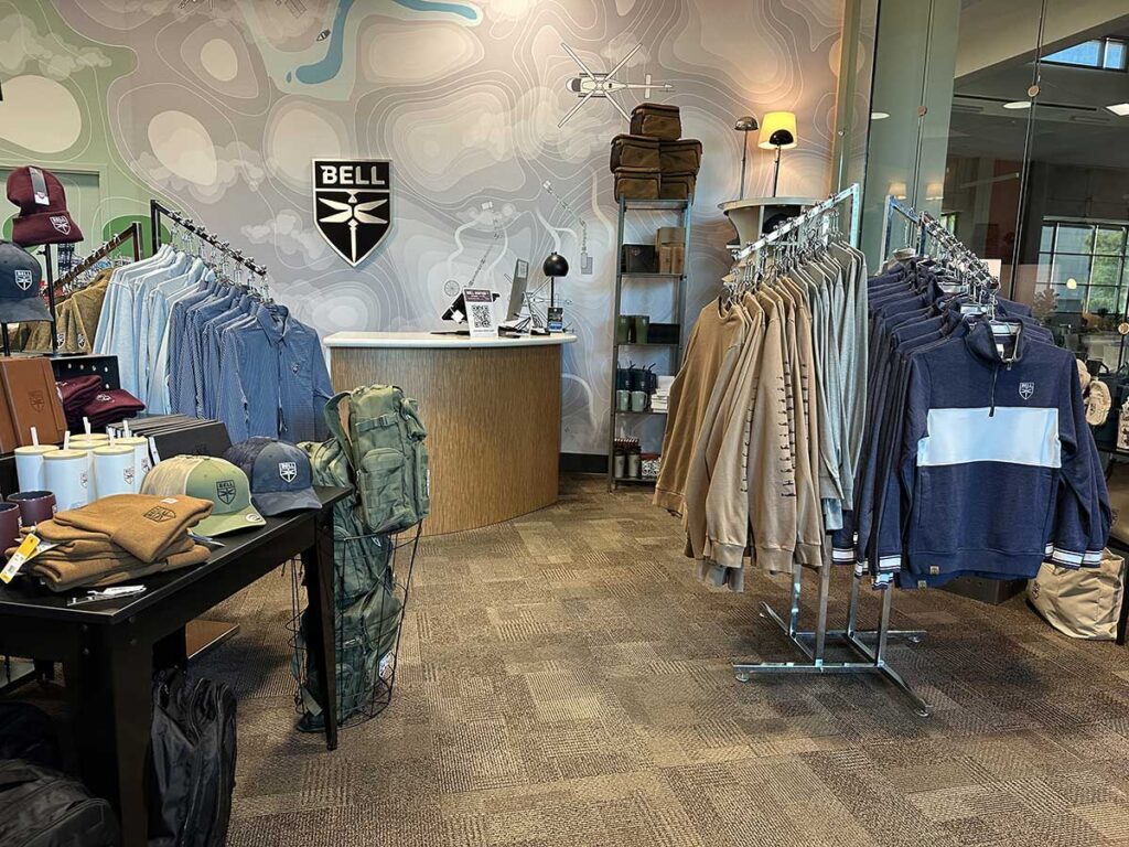 A retail store featuring Bell-branded clothing and accessories. Racks display shirts, jackets, and hats. A checkout counter is in the background with a Bell logo on the wall. Shelves hold various merchandise and apparel.