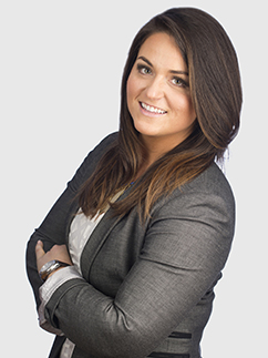 A woman with long brown hair smiles at the camera. She is wearing a grey blazer over a white shirt and has her arms crossed. The background is plain white.