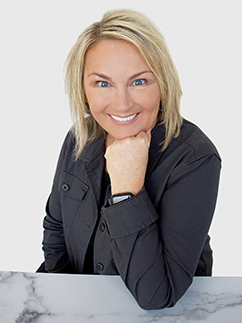 Woman with blonde hair wearing a black jacket smiles at the camera, resting her chin on her hand. She is seated at a marble table with a white background.