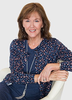 A woman with shoulder-length brown hair is seated on a white chair, smiling. She wears a blue, floral-patterned cardigan over a blue top, with a necklace and bracelets. The background is plain white.