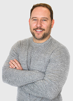 A man with a beard and short hair, wearing a gray sweater, stands smiling with his arms crossed against a light gray background.