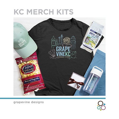 Flat lay of KC merch kit: a black "Grape Vine KC" t-shirt, a blue cap, Velvet Creme popcorn, a water bottle, coffee pack, and a blue cloth. The design features Kansas City landmarks and a cat. Branding by Grapevine Designs.