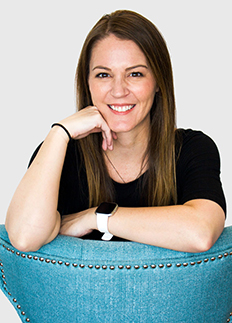 Woman with long brown hair, wearing a black shirt and a smartwatch, smiles while resting her chin on her hand. She leans over a turquoise chair with brass studs, set against a plain background.