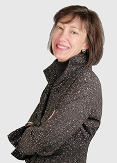 Woman in a speckled black jacket stands against a light gray background, smiling with arms crossed. She has shoulder-length dark hair and is slightly angled to the side.