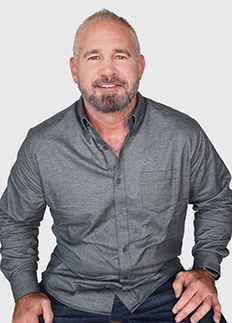 A man with a short beard and gray hair is sitting and smiling, wearing a gray button-up shirt and dark pants against a plain light background.