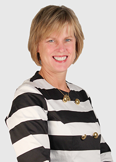 A person with short blonde hair smiles while wearing a black and white striped outfit with decorative buttons. The background is a plain, light color.