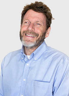 A man with short brown hair and a beard smiles, wearing a light blue dress shirt, against a plain light background.