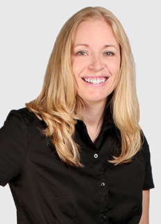 A person with long blonde hair is smiling, wearing a black collared shirt. The background is plain and light-colored.