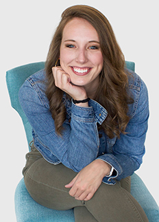 A smiling woman sitting on a turquoise chair, resting her chin on her hand. She has long, wavy brown hair and is wearing a denim jacket and olive green pants. The background is plain white.