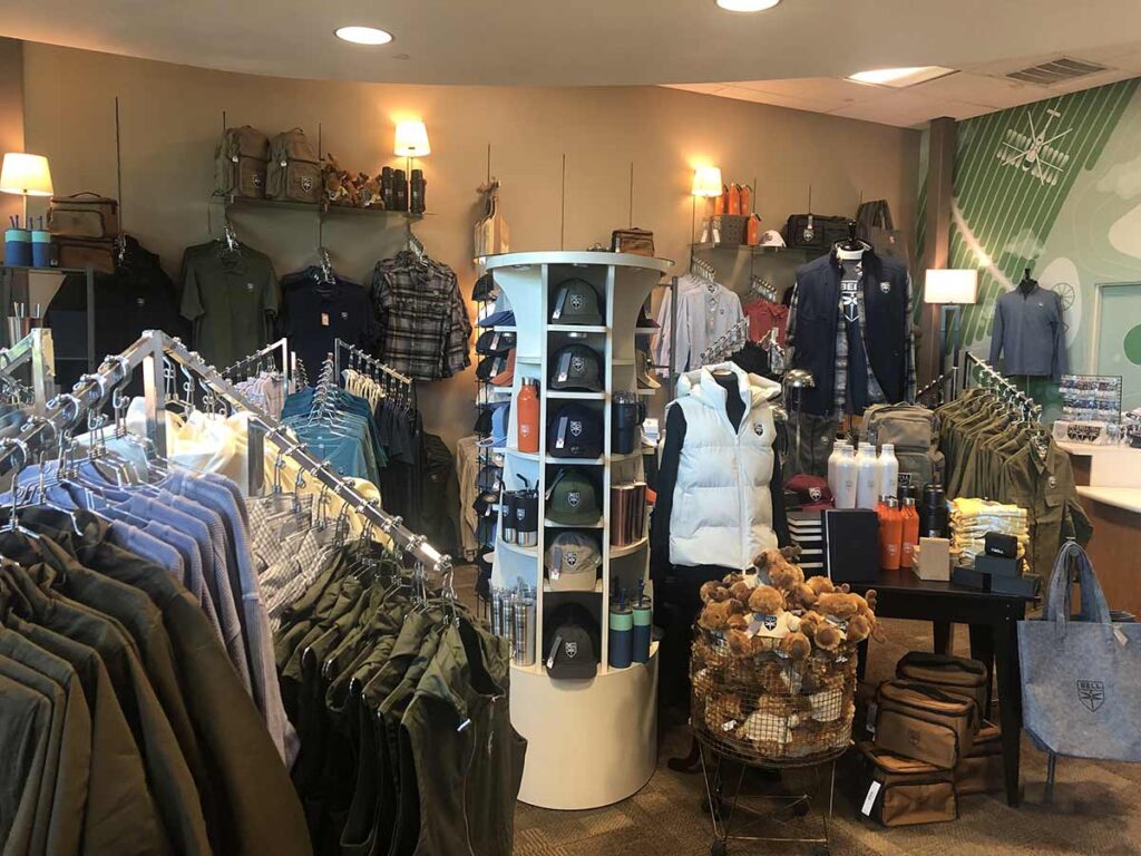 A store display featuring various apparel and accessories. Jackets, hats, and shirts are neatly arranged on racks, with a central shelf displaying water bottles and stuffed toys. The walls are adorned with hanging bags and more clothing items.