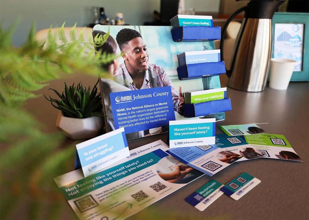 A tabletop display features brochures and cards from NAMI Johnson County, focusing on mental health awareness and support. Brochures are held in blue slots, and additional materials are spread out on the table. A plant and coffee pot are in the background.