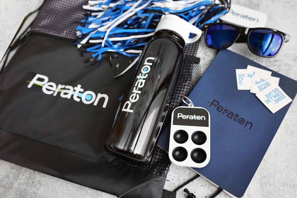 A collection of Peraton-branded items: a black drawstring bag, water bottle, keychain with four buttons, blue notebook, blue sunglasses, and four stickers. Blue and white pom-poms are also visible.