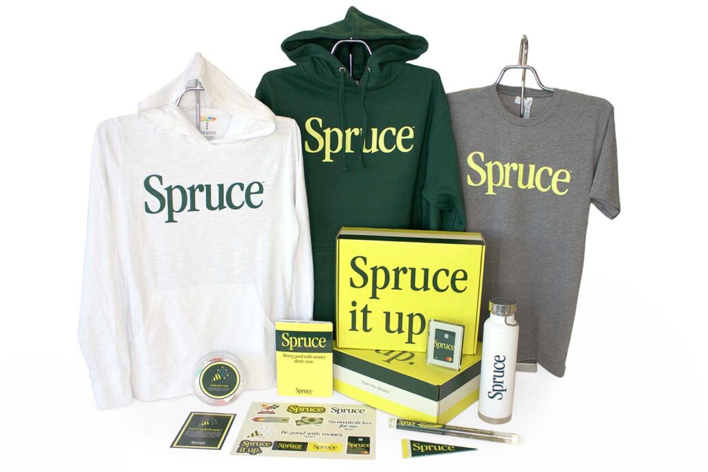 Merchandise display featuring three hoodies and a T-shirt with "Spruce" lettering, alongside a water bottle, stickers, and a box labeled "Spruce it up." Items are in white, green, and gray tones, arranged neatly.