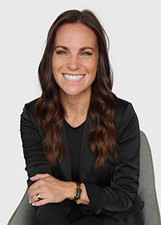 Smiling woman with long, wavy brown hair, wearing a black blazer and a gold watch, sits with arms crossed. She is seated against a plain white background.