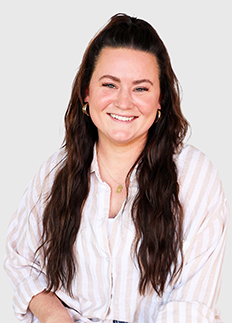 A woman with long brown hair is smiling at the camera. She is wearing a light-colored striped shirt and gold hoop earrings. The background is plain and light-colored.