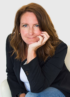 A woman with long brown hair, wearing a black blazer and white shirt, is sitting and resting her chin on her hand. She is smiling slightly and looking directly at the camera, with a neutral gray background behind her.