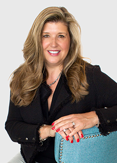 Woman with long blonde hair, wearing a black blazer and red nail polish, smiles while leaning on a turquoise chair with silver studs. She is adorned with a necklace and rings, set against a plain white background.