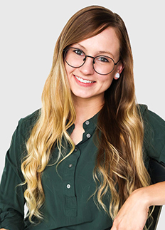 Smiling woman with long wavy blonde hair and glasses, wearing a dark green shirt, sits against a light gray background.