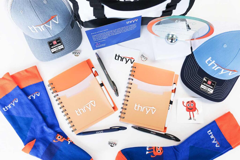 A collection of branded merchandise including two hats, notebooks, socks, pens, a mug, and an award plaque with the "thryv" logo. The items are arranged neatly on a white background.