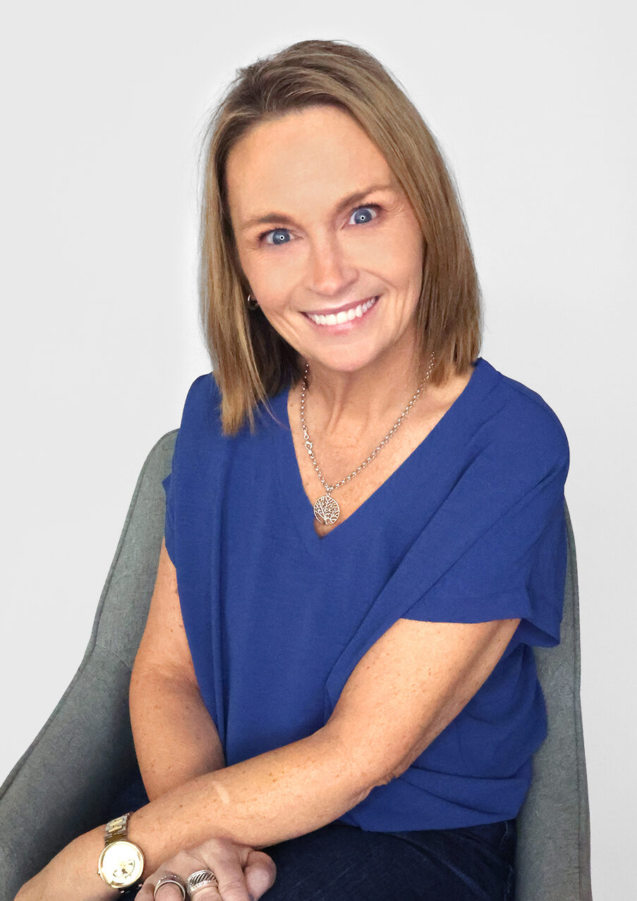A woman with shoulder-length blonde hair, wearing a blue shirt and dark jeans, sits on a gray chair, smiling at the camera. She has blue eyes, a watch, rings, and blue-painted fingernails—perfect for representing a promotional marketing company.