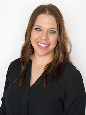 A woman with long brown hair and green eyes, wearing a black blouse, smiles at the camera against a plain white background.