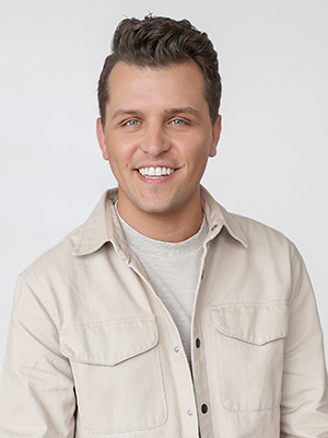 A smiling man with short brown hair wearing a light beige jacket over a light gray shirt, posed in front of a plain white background.