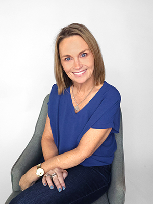 A woman with shoulder-length light brown hair sits on a gray chair, smiling at the camera. She wears a blue short-sleeve top, dark jeans, a gold necklace, and a watch. The background is plain white.
